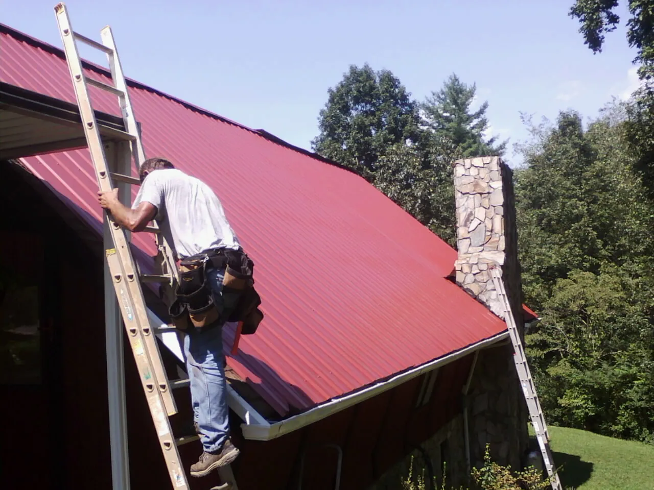 Roofer on a ladder beside a red metal roof and stone chimney — Soffit & Fascia Repair by Roofing Brothers in Frenchtown